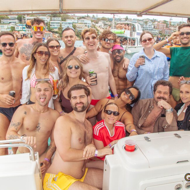 A group of people in swimwear and sunglasses pose together on a boat, smiling and having fun. There are drinks in hand, water in the background, and waterfront buildings visible under a partly cloudy sky.