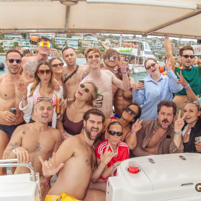 A group of people in swimsuits and summer attire smile and pose together on a boat, with blue water and buildings in the background. Some hold drinks, and the atmosphere is lively and festive.