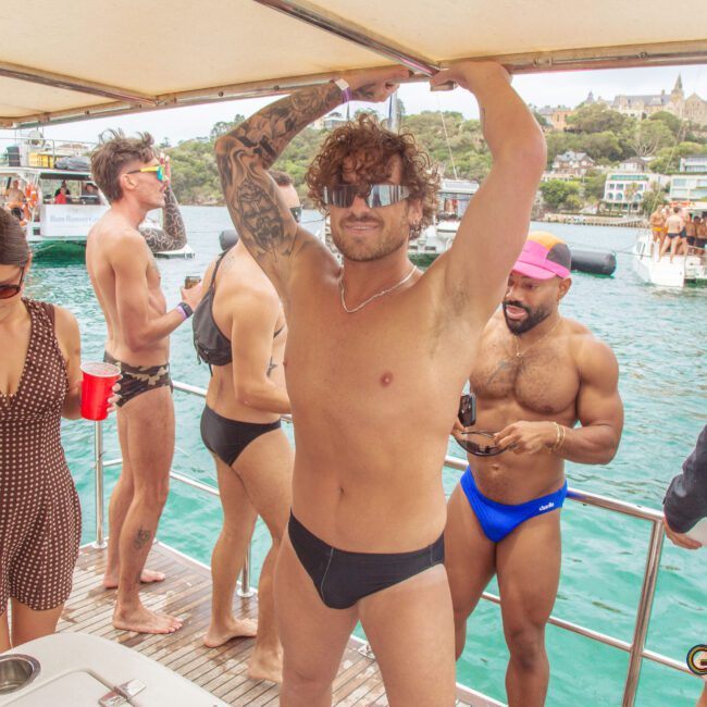 A group of people in swimwear party on a boat. A man in sunglasses and black swim briefs poses at the center, with others socializing and holding drinks. Water, boats, and buildings are visible in the background.