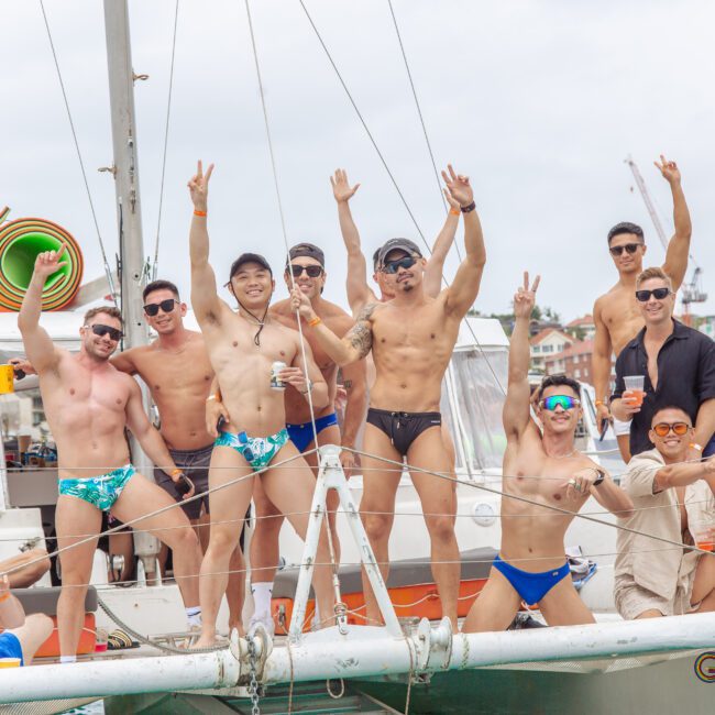 A group of men in swimwear pose and smile on a sailboat, some holding drinks and making peace signs, with houses visible in the background under a cloudy sky.