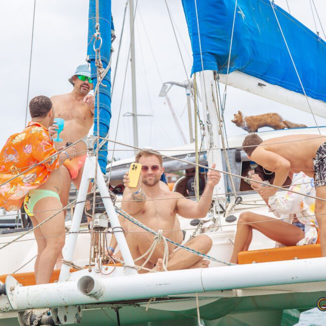 Four men in swimwear relax on the deck of a sailboat, with one holding a drink and another wearing a colorful shirt and bucket hat. A cat stands on the boat in the background. The atmosphere is fun and casual.