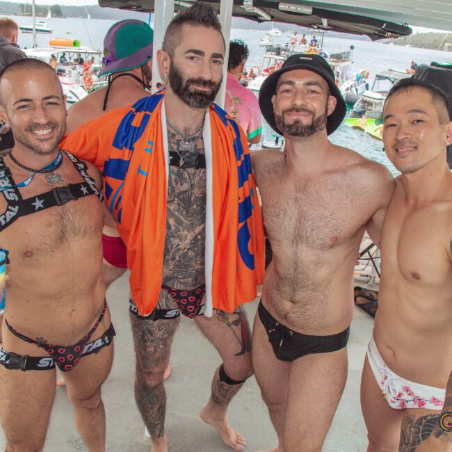 Four smiling men in swimwear pose together on a boat at a lively party, with others and boats visible on the water in the background. The atmosphere is festive and sunny.