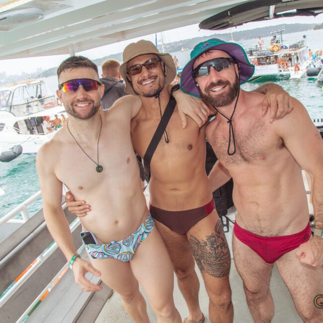 Three smiling men in colorful swim briefs and hats pose together on a boat, with water, other boats, and people in the background at a lively outdoor event.