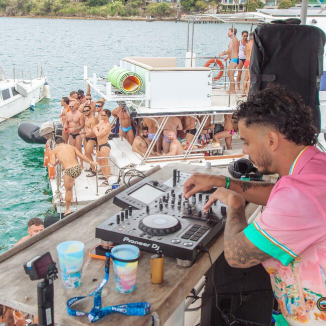 A DJ in a pink shirt mixes music on a deck overlooking a lively boat party, with people swimming, dancing, and socializing on several boats in a turquoise bay.