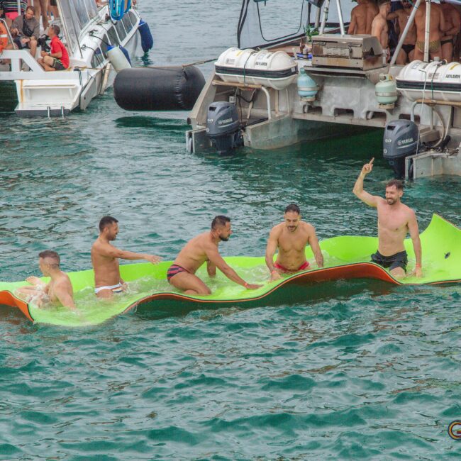 Five men in swimwear are sitting and standing on a large green floating mat in the water near anchored boats. One man is standing and raising his hand, while the others are sitting or kneeling and smiling.