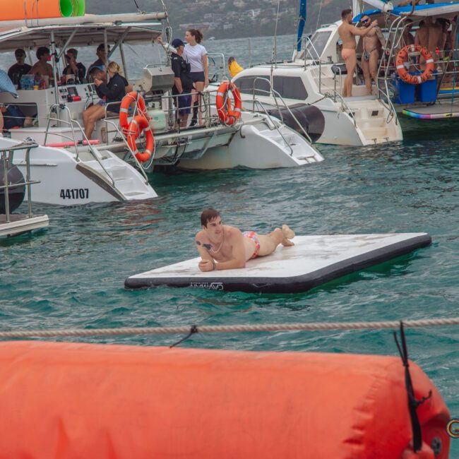 A person in a swimsuit lies on a floating mat in the water near several anchored boats with people onboard. The scene looks festive and social, set in a bay with hills in the background.