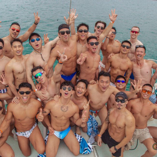 A group of shirtless men in swimwear pose together on a boat, smiling and making peace signs, with blue water visible in the background.