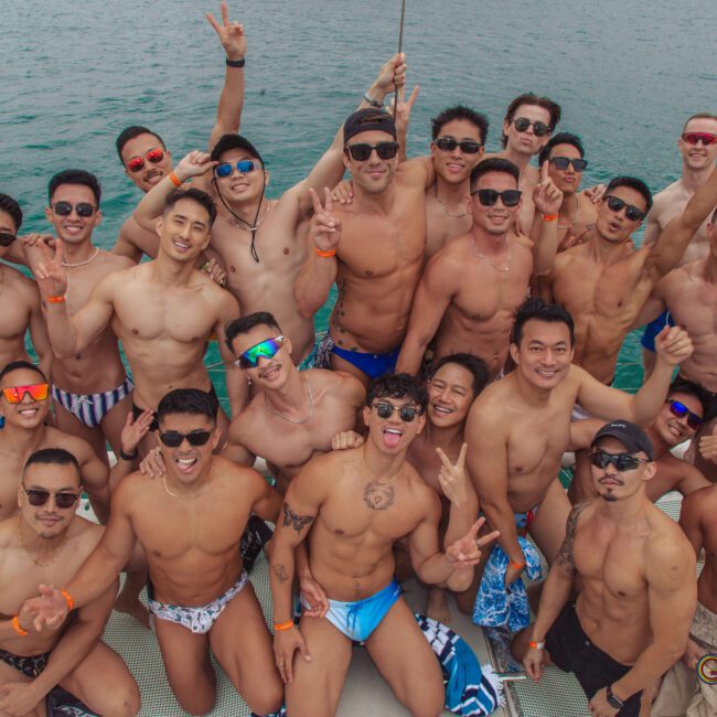 A group of shirtless men in swimwear pose and smile on a boat, many holding drinks and flashing peace signs, with blue water in the background and a festive, joyful atmosphere.