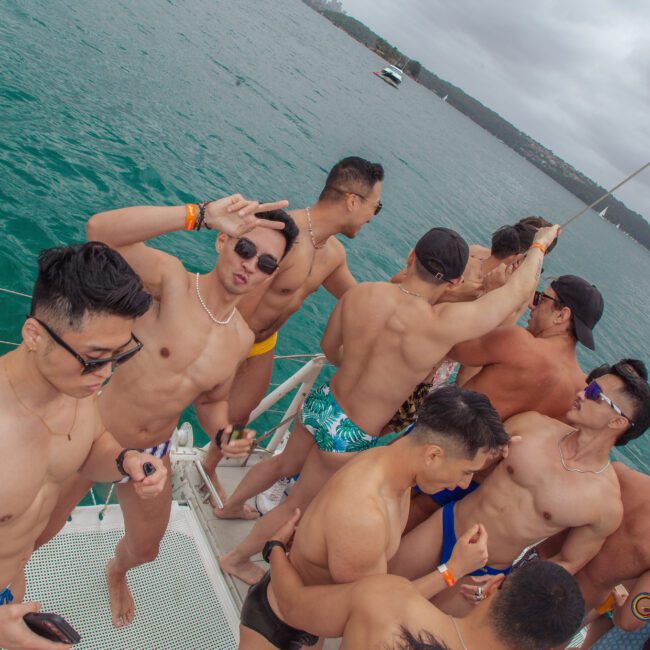 A group of shirtless men in swim trunks are dancing and socializing on a boat deck, with the ocean and boats visible in the background under a cloudy sky.