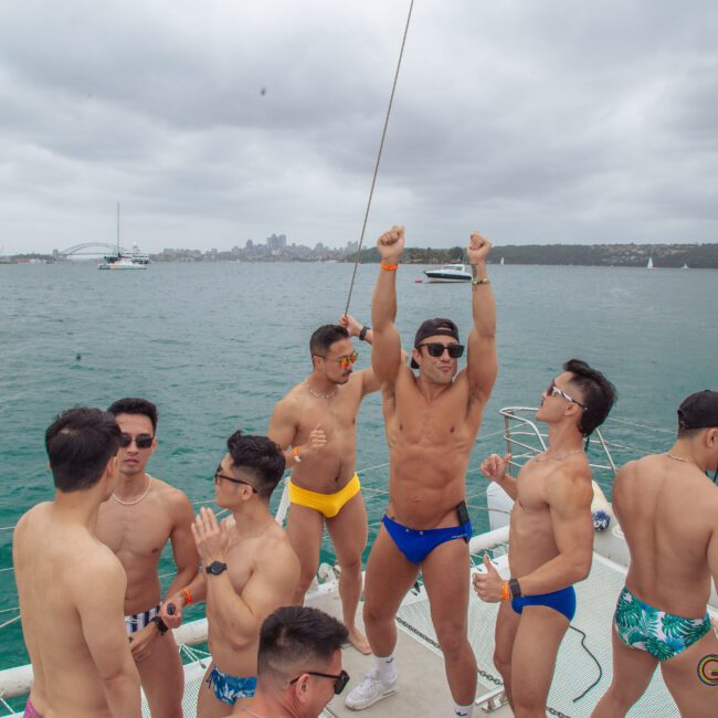 A group of men in colorful swimwear are dancing and socializing on a boat, with a city skyline and cloudy sky in the background on the water.