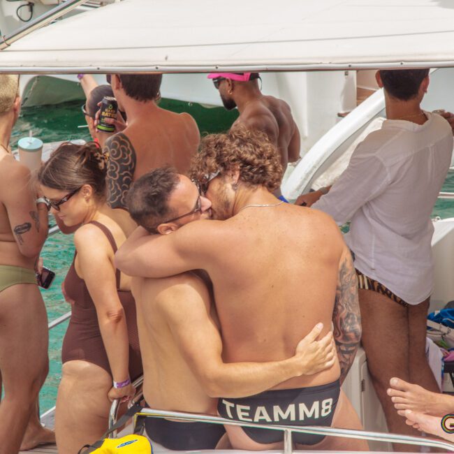 Two men embrace and kiss on a boat crowded with people in swimwear, enjoying a sunny day on the water. Others are chatting, holding drinks, and relaxing as the boat floats on turquoise water.