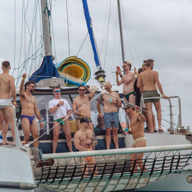 A group of men in swimwear are standing and socializing on the deck of a boat named "Ocean Life" on a cloudy day, with some holding drinks and others chatting or looking towards the water.