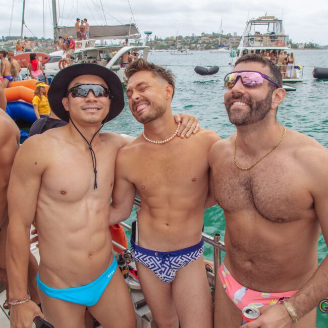 Three men in colorful swimwear smile and pose together on a boat during a lively party on the water, with other people, boats, and cityscape visible in the background.