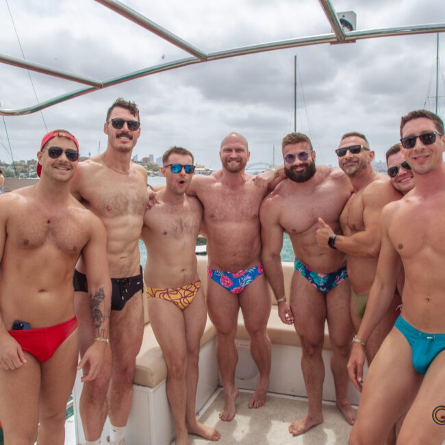 A group of eight men in colorful swim briefs pose together on a boat, smiling at the camera. Other boats and people are visible in the background on the water under a cloudy sky.