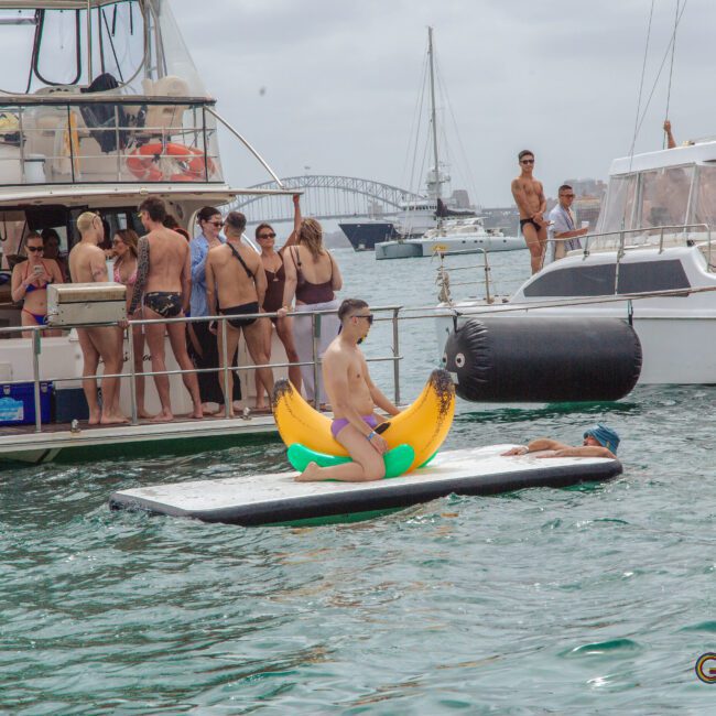 A man sits on an inflatable banana float in the water near two boats with groups of people socializing and relaxing. The Sydney Harbour Bridge is visible in the background under a cloudy sky.