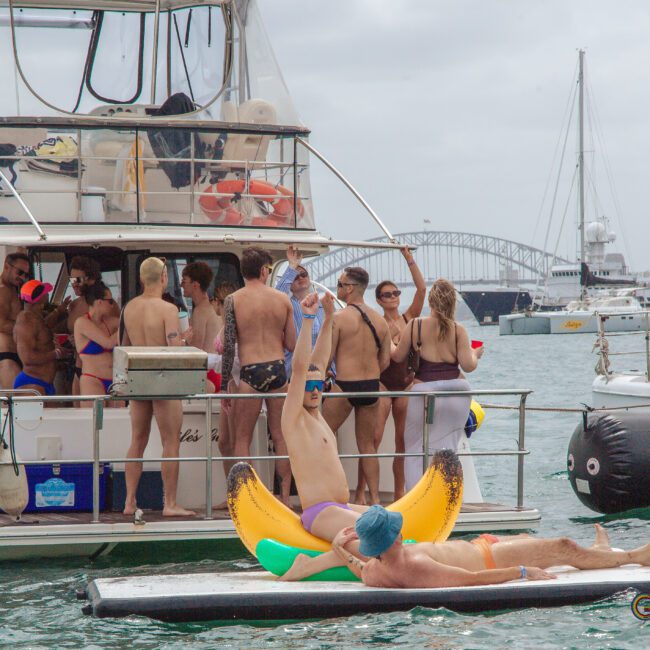 A group of people in swimsuits gather and socialize on a boat, while two people float on pool inflatables and a paddleboard beside the boat. Other boats and a large bridge are visible in the background.