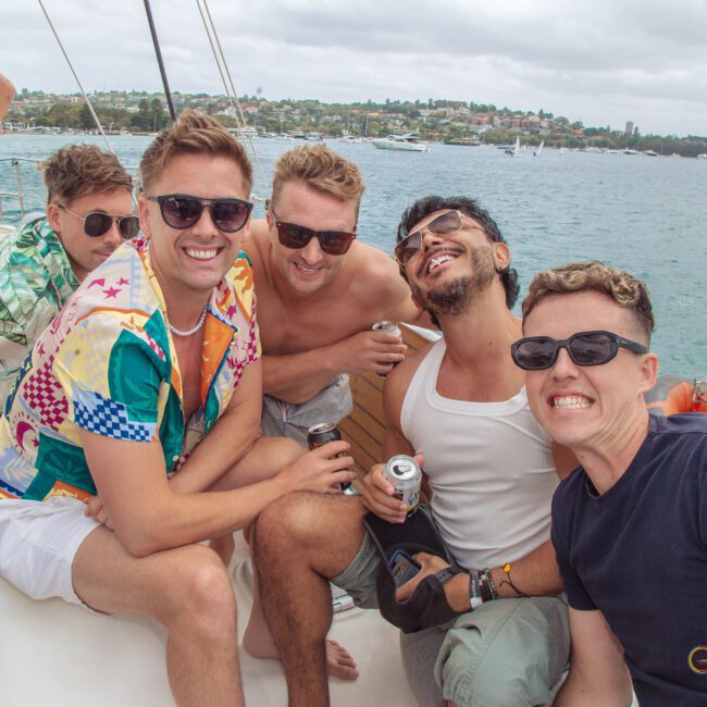 A group of five men smile and pose together on a boat, holding drinks. Other people are in the background, and the boat is on the water with a city and greenery visible across the bay.