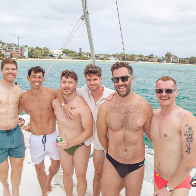 Six men in swimwear stand smiling together on a boat, with blue water and a coastal town in the background. The weather is cloudy but bright. The logo “GAYM” appears in the bottom right corner.
