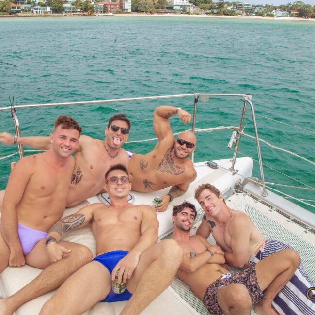 Six young men in swimwear relax and pose happily on the deck of a boat, with turquoise water and a shoreline with houses visible in the background. Some smile and make playful gestures. The CAYM logo is in the corner.