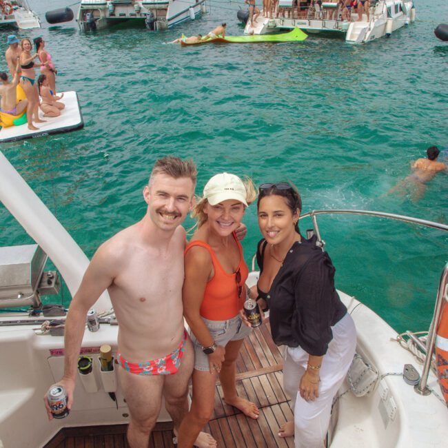 Three people stand smiling on a boat, holding drinks, with several other boats and groups of people partying in the turquoise water behind them on a cloudy day. The atmosphere is festive and summery.