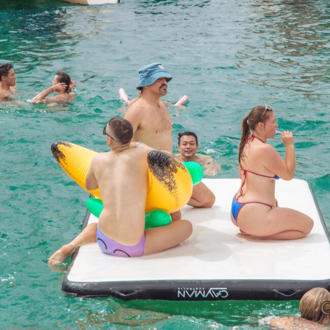 A group of people in swimsuits relax and play on a floating mat in clear, turquoise water. Some are sitting or lying on the mat, while others swim around it, enjoying a sunny day.