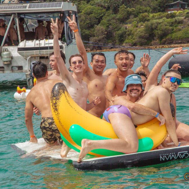 A group of smiling men in swimsuits pose together on an inflatable banana float in the water, waving and making peace signs. Other people and a docked boat are visible in the background on a sunny day.