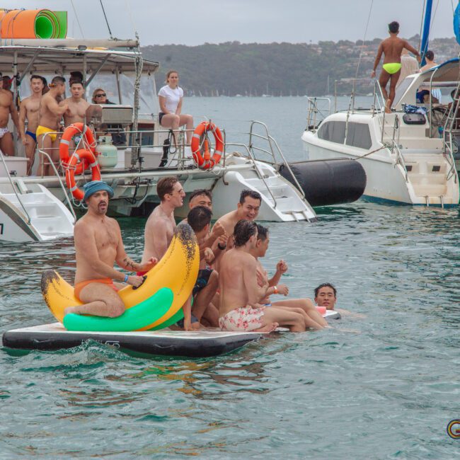 A group of people in swimwear laughing and playing on a banana-shaped inflatable float in the water, with boats and more people in the background. It looks like a lively, festive gathering on a cloudy day.