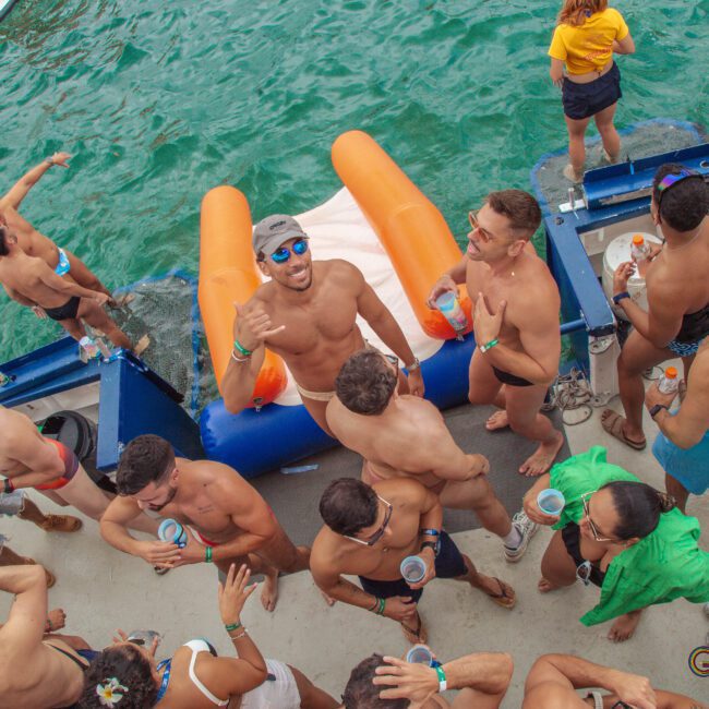 A group of men in swimwear socialize, drink, and smile on a dock by the water with an inflatable float nearby during a daytime pool or boat party.