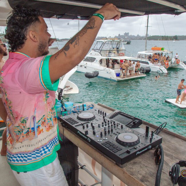 A DJ in a colorful shirt plays music on a boat while people in swimwear dance and relax on nearby boats and a floating platform in a lively outdoor party scene on the water.