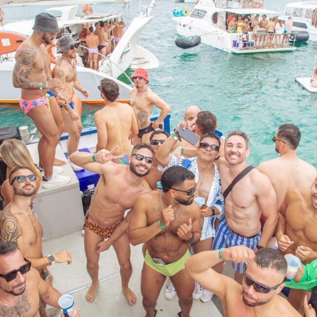 A lively crowd of men in swimwear pose and dance on a dock beside boats at a festive outdoor party on the water, with others mingling on nearby boats under a cloudy sky.
