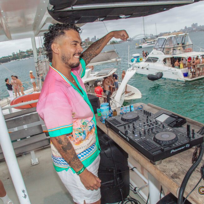 A smiling DJ in a colorful shirt performs on a boat deck with DJ equipment, while people in swimwear dance and relax on nearby boats and in the water under a cloudy sky.