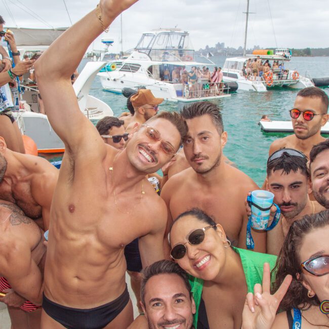 A group of people in swimwear smile and pose for a selfie on a boat, with other boats and people in the background on the water. The mood is festive and sunny.