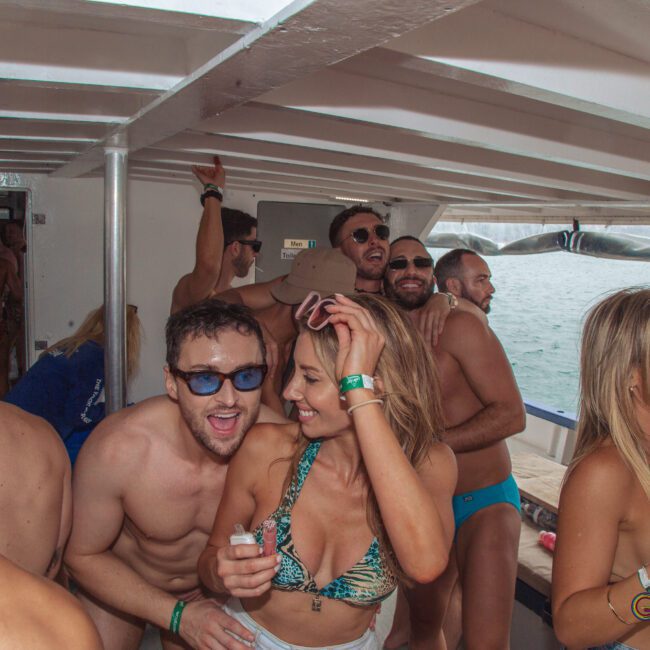 A group of people in swimwear enjoy a lively party on a boat. Some are dancing and smiling, while others are holding drinks. The atmosphere appears festive and carefree, with the sea visible in the background.