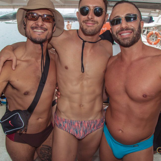 Three men in swimsuits smile and pose together on a boat, arms around each other, with other people and water visible in the background. They appear to be enjoying a sunny day at a pool or beach party.