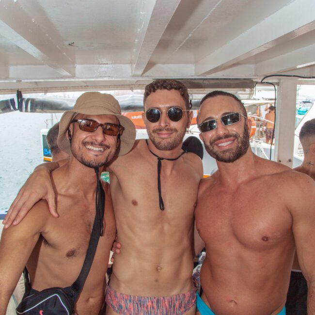 Three men in swimsuits and sunglasses stand close together and smile under a shaded area, with bright outdoor light in the background. Two wear hats, and the scene has a lively, beach or pool party atmosphere.