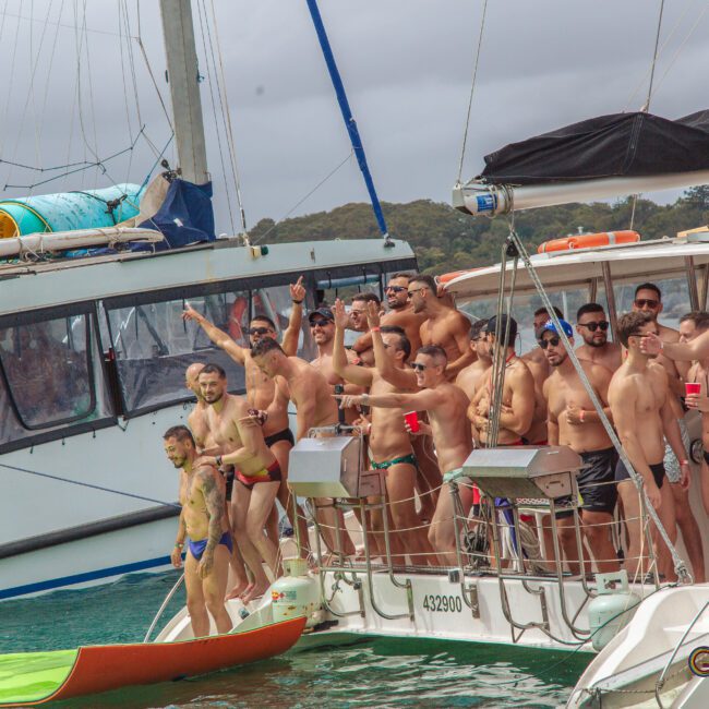 A group of men in swimwear stand and pose on the decks of two docked boats, many smiling and waving at the camera, with overcast skies and water in the background.