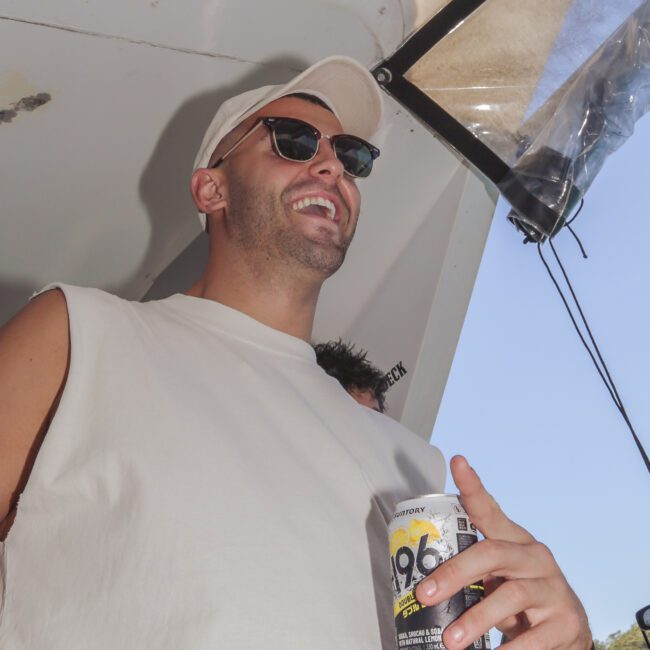 A man wearing sunglasses, a white cap, and a sleeveless white shirt smiles while holding a can of Suntory 196. He is standing under a boat canopy with daylight and trees visible in the background.