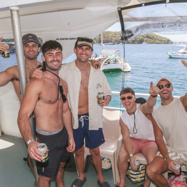 Five men smiling and posing together on a boat, holding drinks. They are dressed in swimwear and sunglasses, with water, boats, and greenery visible in the background on a sunny day.