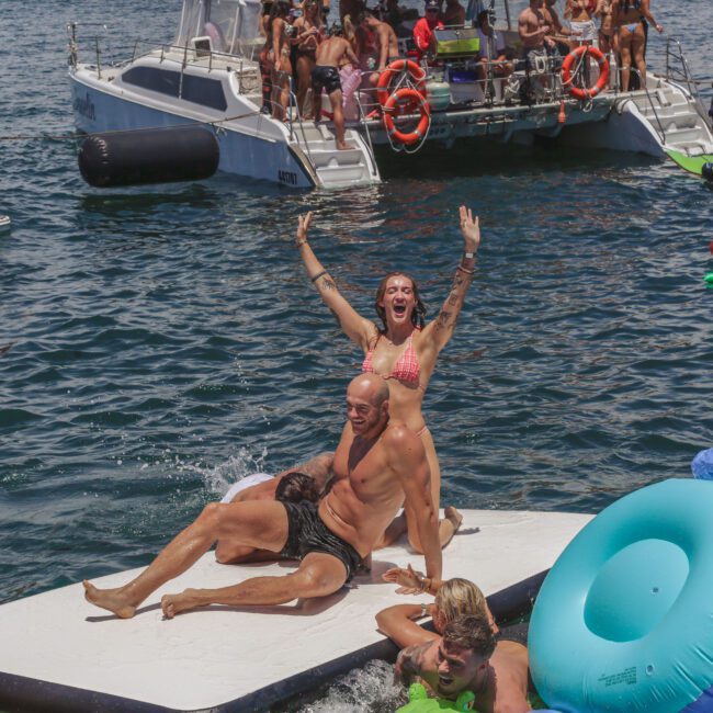 People enjoy a lively party on the water, with individuals playing and laughing on a floating mat, surrounded by colorful inflatables and two boats in the background packed with people.