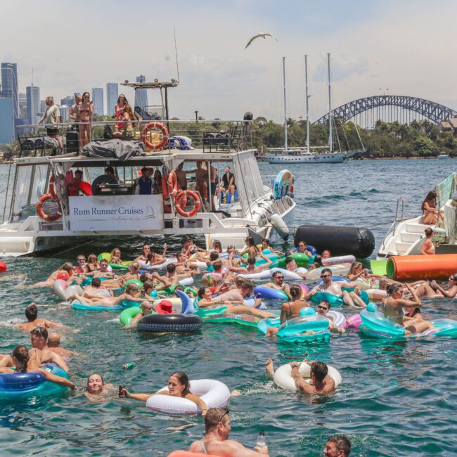A large group of people on colorful inflatables float in the water near boats with "Rum Runner Cruises" signage, with a city skyline and Sydney Harbour Bridge visible in the background.