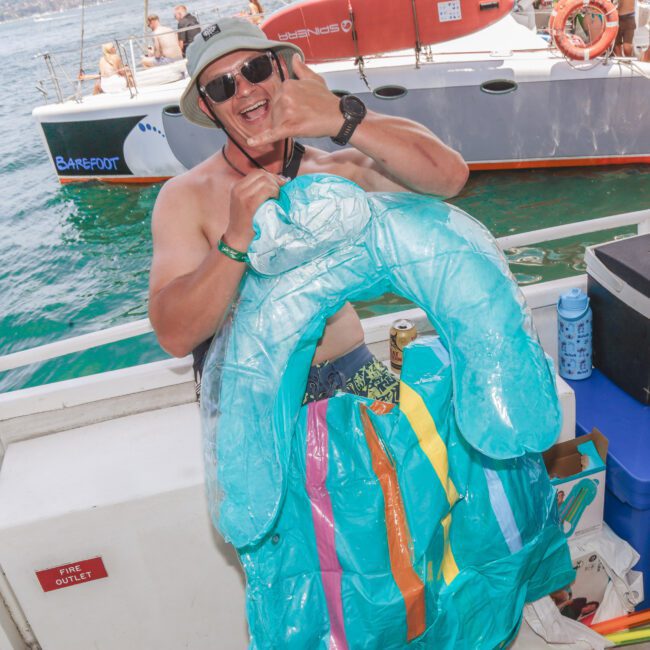 A smiling man in sunglasses and a bucket hat stands on a boat holding a deflated blue inflatable pool float, giving a shaka hand sign. The sea and another boat are visible in the background.