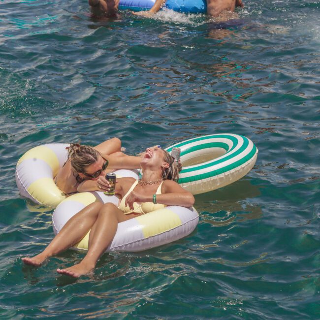 Two women relax and laugh on an inflatable pool float in the water, enjoying a sunny day. Other people float and swim around them. The scene is lively and joyful.