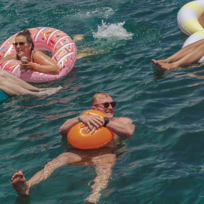 Several people relax in the water on colorful inflatable pool floats, smiling and enjoying drinks on a sunny day. The water is clear and blue, and everyone appears to be having fun.