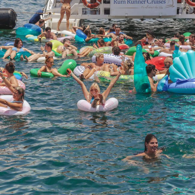 A large group of people float on colorful inflatable rafts and pool toys in the water near a boat, enjoying a sunny day with smiles and raised arms. The boat has "Rum Runner Cruises" written on it.
