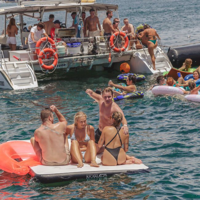 A group of people enjoy a party on the water, lounging on floats and swimming near a boat. Some are sitting on an inflatable mat, while others are in the sea or on the boat. The weather is sunny and festive.