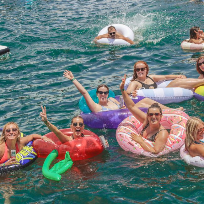 A group of smiling adults relax on colorful pool floats in a lake, waving and enjoying the sunshine. The water is clear and blue, and everyone looks cheerful and festive. The "Yacht Social Club" logo is visible in the corner.