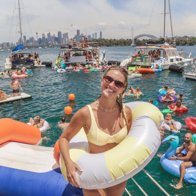 A smiling woman in a bikini with pool floats stands on a boat, surrounded by people relaxing on colorful inflatables in the water. Several boats and a city skyline, including the Sydney Harbour Bridge, are in the background.