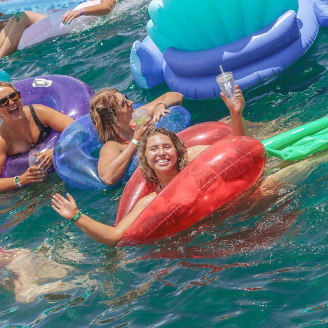 A group of smiling women float on colorful inflatable pool floats in clear blue water. One woman in the center waves and holds a drink, enjoying a sunny day. The mood is joyful and festive.