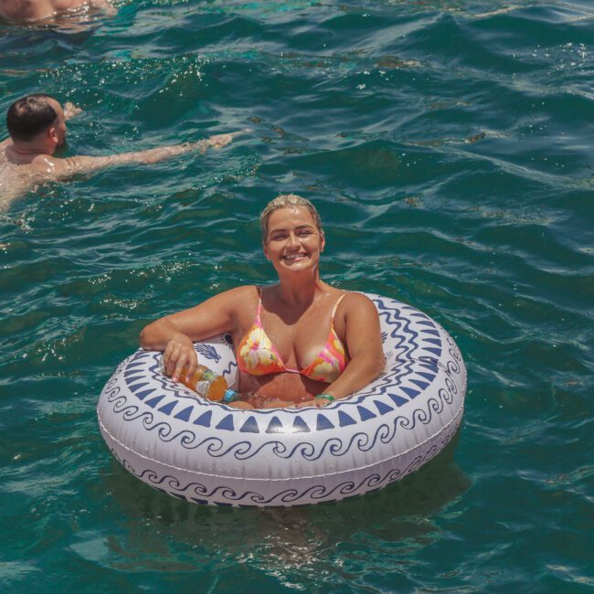 A smiling woman in a bikini sits in a decorative inflatable ring on bright blue water while holding a drink, with other people swimming nearby. The words "Yacht Social Club" are visible in the bottom right corner.
