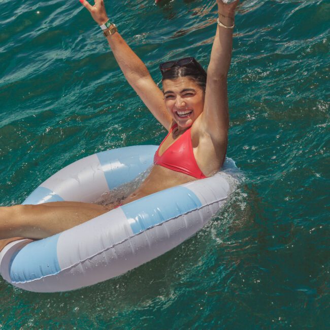 A smiling woman in a red bikini floats on a blue-and-white inflatable ring in the sea, raising her arms joyfully. Sunlight sparkles on the turquoise water. A "Yacht Social Club" logo appears in the bottom right corner.
